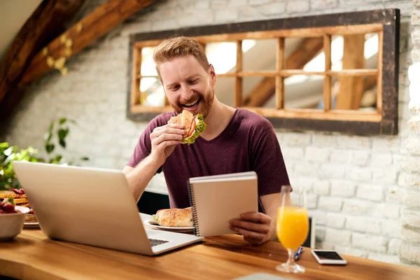 Happy man eating breakfast while reading notes and working at home.