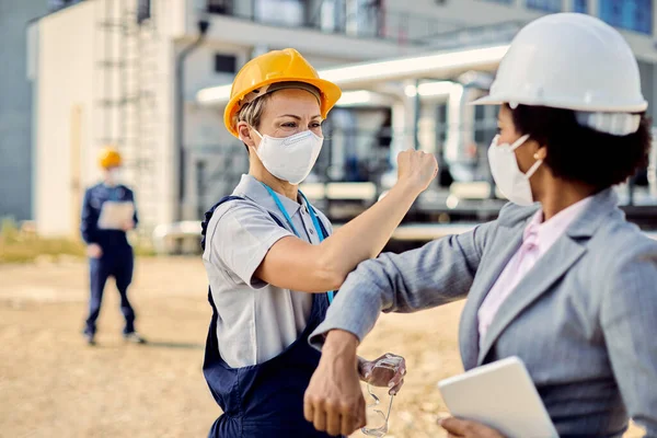 Female civil engineer and building contractor greeting with elbow while wearing protective face masks at construction site.