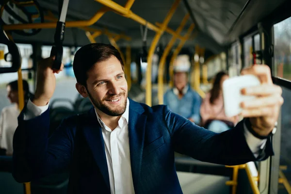 Happy male entrepreneur using smart phone and taking selfie while commuting by bus.