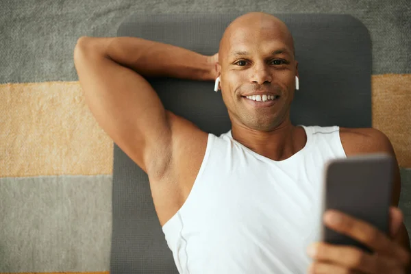 Happy African American athletic man listening music over earphones and using mobile phone while lying down on floor after home workout.