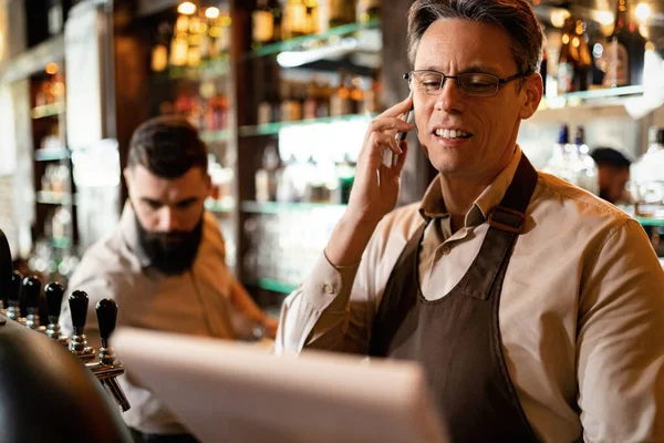 Smiling bartender talking on mobile phone while doing paperwork in a pub.