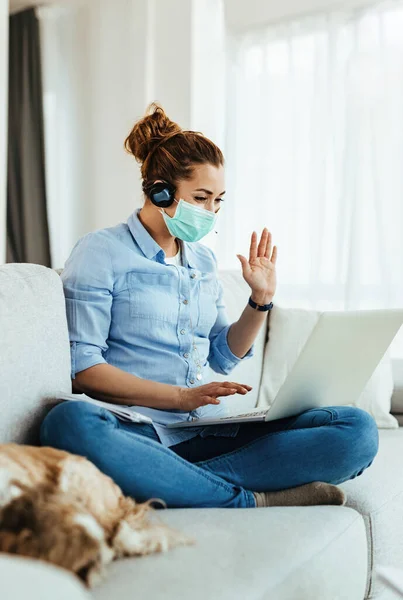 Happy woman with protective face mask using laptop and waving to someone while making video call from home.