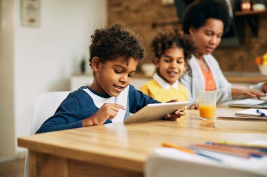 Happy African American boy using touchpad while relaxing at home. His mother and sister are in the background. 