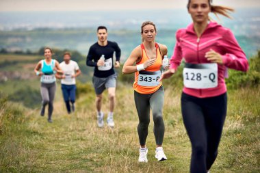 Group of runners taking a part in marathon race in nature. Focus is on woman in orange shirt. 