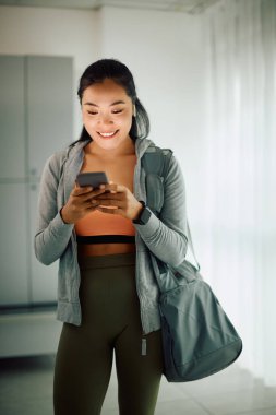 Young happy Asian athletic woman texting on smart phone at gym's dressing room.