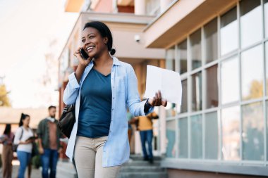 Happy African American university student holding test results and communicating over smart phone at campus.