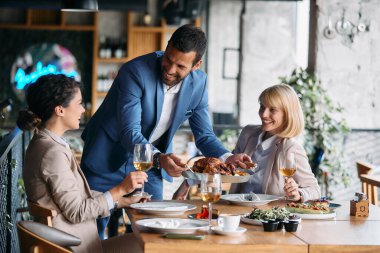 Happy businesswomen having fun while their male colleagues is serving them lunch in a restaurant. Focus is on businessman. 
