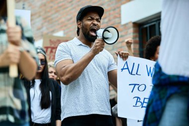 Displeased African American man shouting through megaphone while demonstrating for human rights with crowd of people on the streets. 