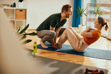 Athletic couple exercising together at home. Happy man is helping his wife while she is practicing sit-ups on the floor. 