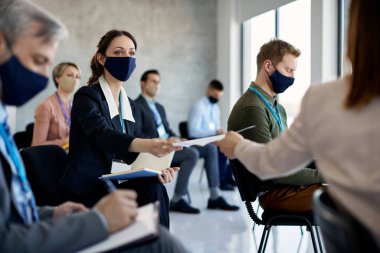 Female entrepreneur wearing protective face mask while taking paper for a colleague during business education event in conference hall.