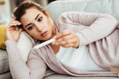 Young woman looking at thermometer in disbelief while measuring her body temperature at home. 