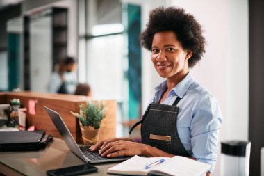 Female African American hairstylist suing laptop and looking at camera while working at hair salon. 