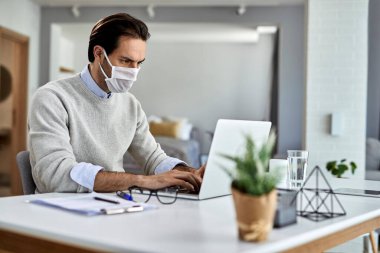 Freelance worker using laptop and wearing protective face mask while working at home during coronavirus epidemic. 