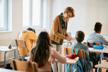 Mature teacher talking to schoolboy while assisting him during a class in the classroom.