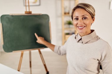 Happy female professor teaching from home and pointing at blackboard while looking at camera. 
