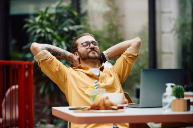Young man with hands behind head relaxing after using laptop in a cafe.