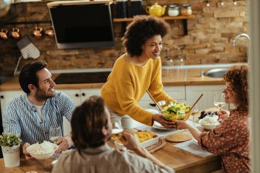 Group of happy friends enjoying while having lunch together at dining table. Focus is on African American woman giving salad to her female friend. 