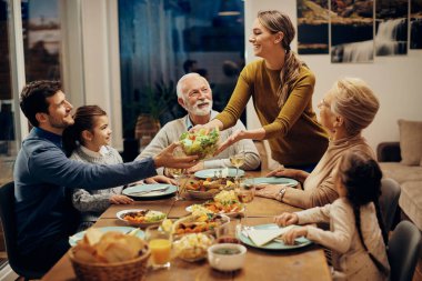 Happy multi-generation family enjoying in a lunch together at home. Focus is on young woman serving salad at dining table. 