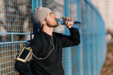 Young male runner drinking water from a bottle while leaning on a fence outdoors. 