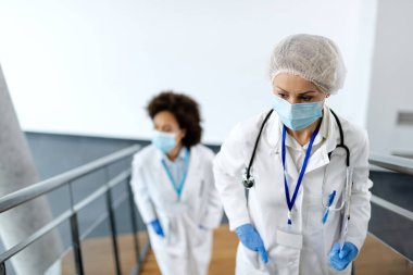 Worried female doctor wearing protective face mask and walking upstairs in a hallway at medical clinic. Her colleague is in the background. 