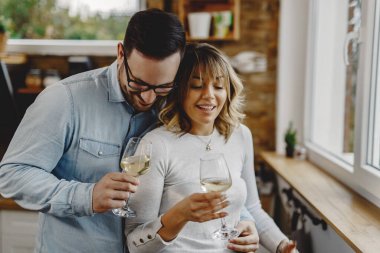 Happy man embracing his wife while drinking wine together in the kitchen. 