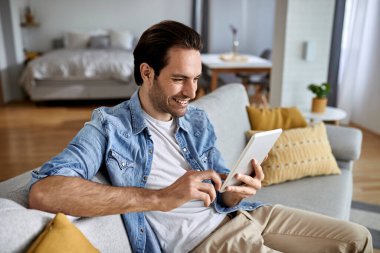 Young happy man surfing the net on touchpad while relaxing at home. 