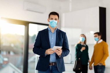 Male real estate agent with digital tablet looking at camera while wearing protective face mask. There is a couple in the background. 