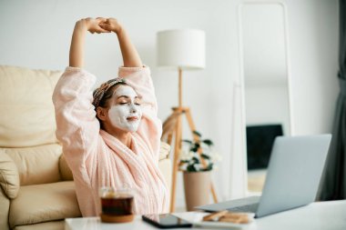 Bornozlu genç bir kadın yüz maskesi takıyor ve evde laptopla çalıştıktan sonra geriniyor.. 