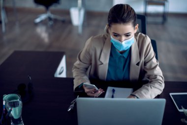 Busy female entrepreneur working on a computer while using cell phone at her office desk. She is wearing protective face mask due to COVID-19 pandemic. 