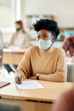 African American female student taking notes at lecture hall and wearing protective face mask due to COVID-19 pandemic.