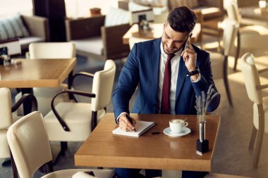  Happy businessman talking on cell phone and writing in notebook while sitting in a cafe. 