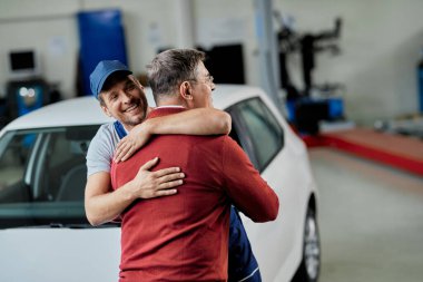 Happy auto repairman and his customer embracing while greeting in a workshop. 