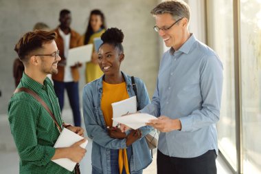 Happy students talking with their professor while standing at university hallway. Focus is on African American student. 