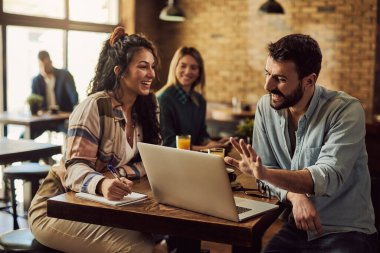 Happy man using laptop and talking to his female friend who is taking notes in a cafe. 