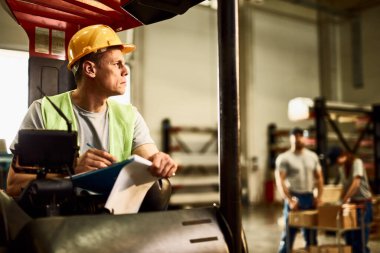 Pensive forklift operator writing on clipboard while working in industrial warehouse. 