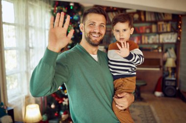 Happy father and his small boy greeting someone during video call on Christmas and looking at camera.