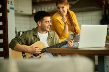 Happy man having fun and playing acoustic guitar while his wife is singing at home.