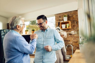 Happy adult son and his mature father holding hands while greeting each other in the kitchen. 