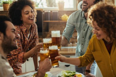 Happy African American woman and her friends having fun while toasting with beer in dining room. 
