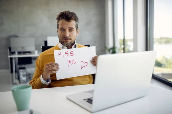 Businessman holding a placard with \'miss you\' inscription while having video call over laptop in the office.