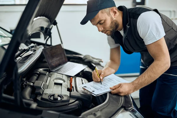 Young mechanic filling paperwork while examining car engine at auto repair shop.