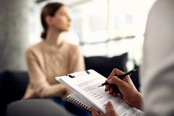 Close-up of psychologist having appointment with female patient and writing notes during psychotherapy.