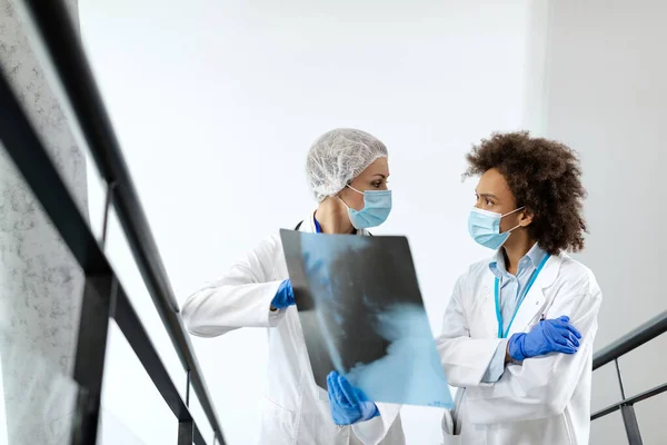 Female radiologists discussing while analyzing patient\'s X-ray image at hospital hallway. They are wearing protective face masks due to coronavirus pandemic.