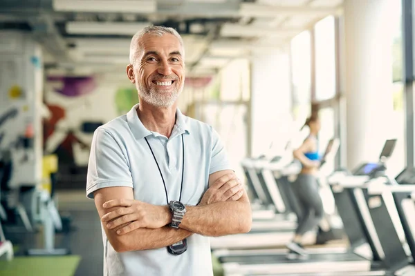 Happy mature personal trainer stands with arms crossed in gym an