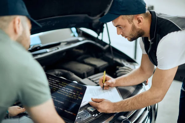 Young mechanic writing notes while examining car engine with a coworker who is using laptop in a workshop.