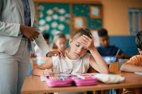 Elementary student holding her head in pain while teacher is consoling her on a class at school. 