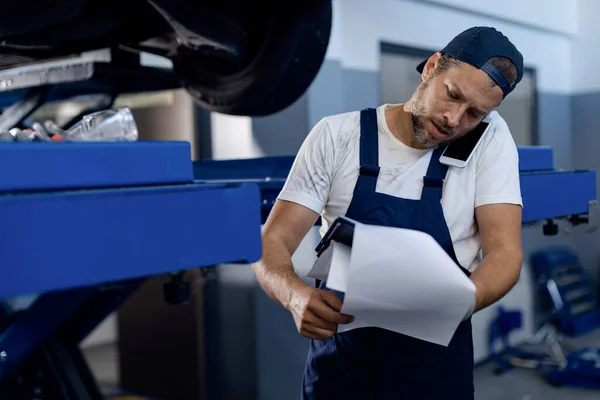 Auto mechanic examining paperwork and communicating over mobile phone in a repair shop.