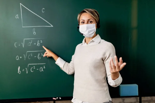 Female teacher with a face mask standing in front of chalkboard and giving online math lecture during coronavirus epidemic.