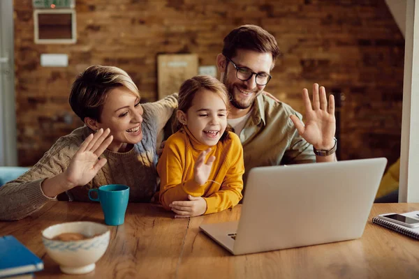 Happy parents with daughter greeting someone while having video call over laptop at home.