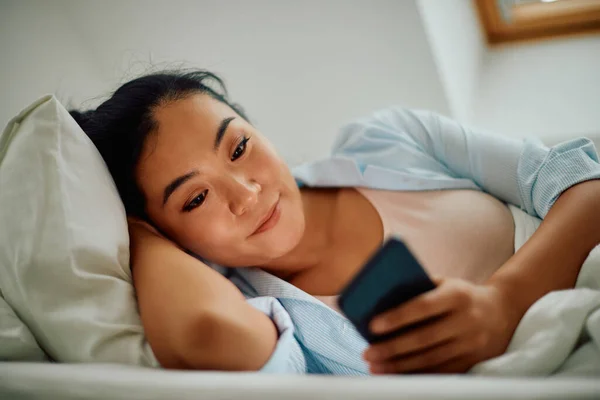 Young Smiling Asian Woman Texting Mobile Phone While Lying Bed — 图库照片